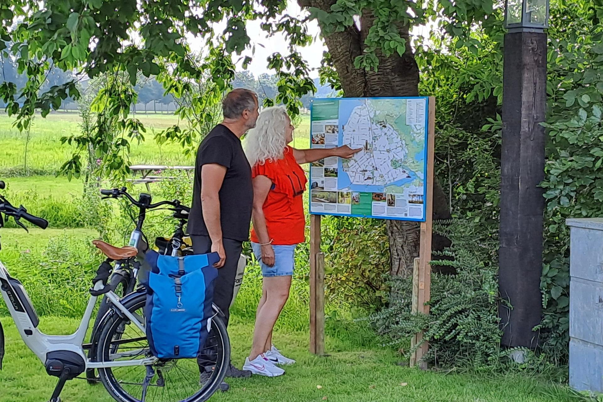 Twee mensen staan voor een informatiebord te kijken. Een persoon wijst iets aan op het bord. Naast hen 2 fietsen met tassen erop.